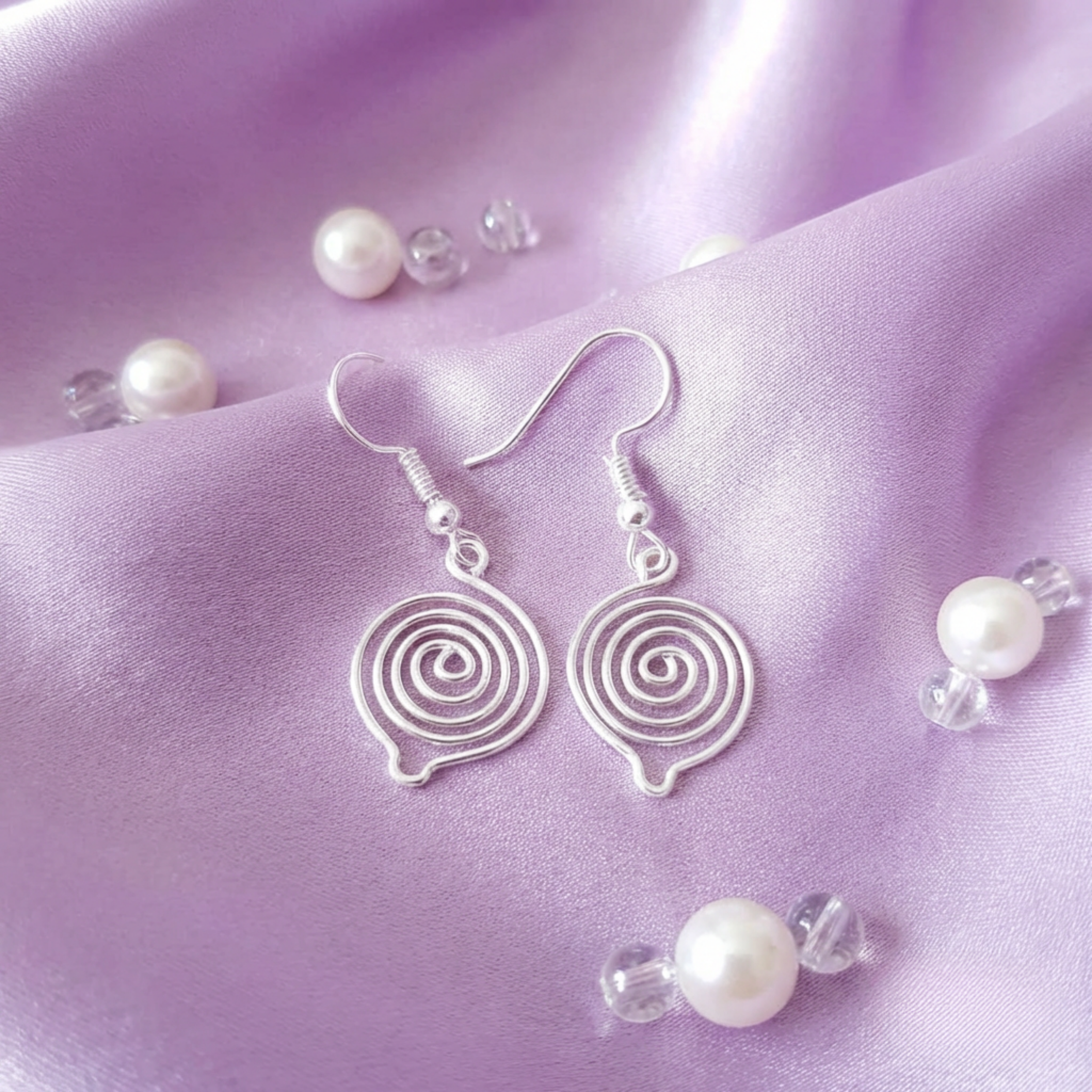 Silver spiral earrings on a purple fabric background with scattered pearls and crystals.