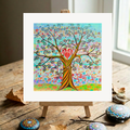 Colourful tree of life painting on a small easel with decorative stones and twigs on a wooden surface.