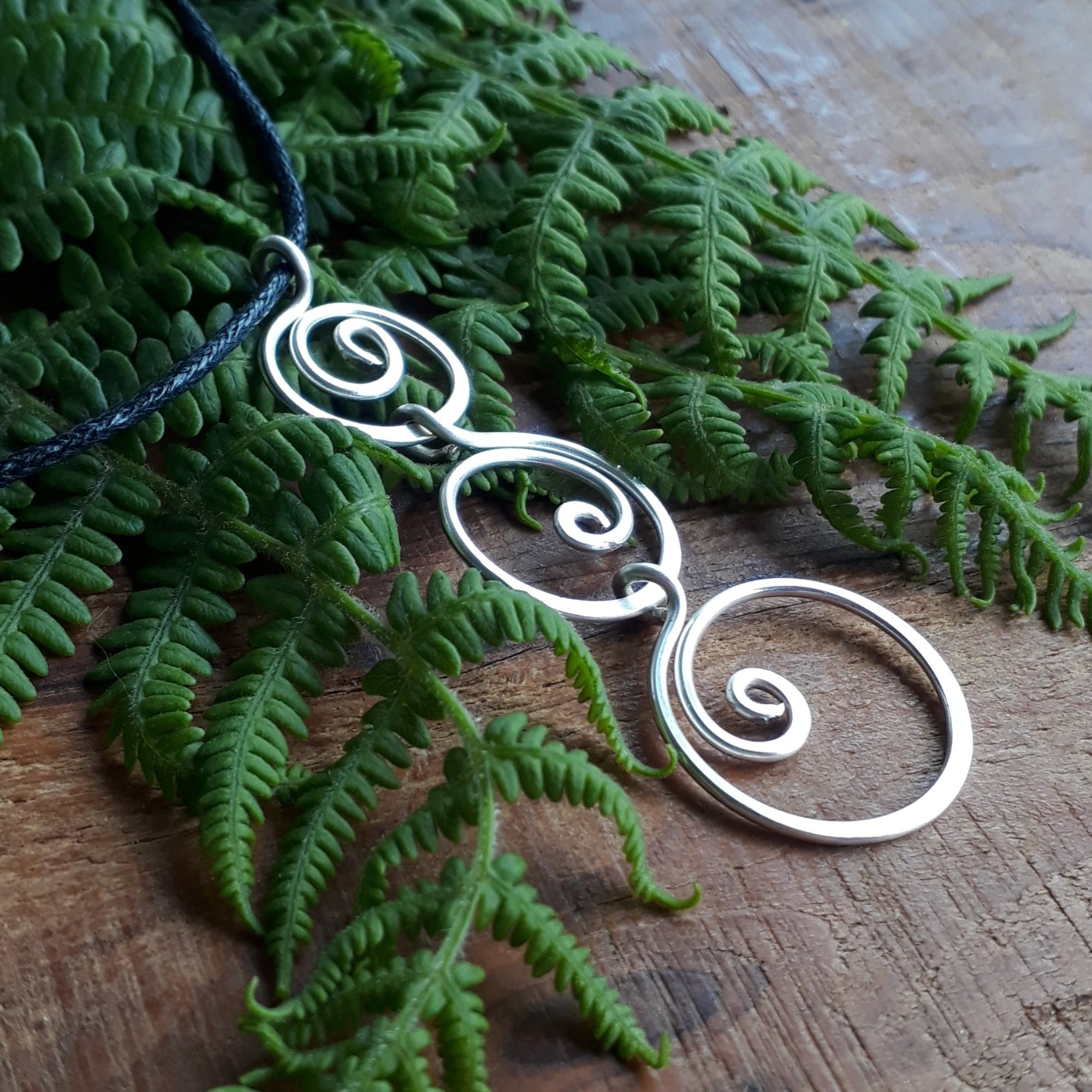 Silver spiral pendant on a dark cord with green fern leaves on a wooden surface