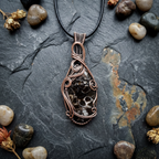 Copper wire-wrapped pendant with embedded stones on a dark stone surface with pebbles and dried plants.