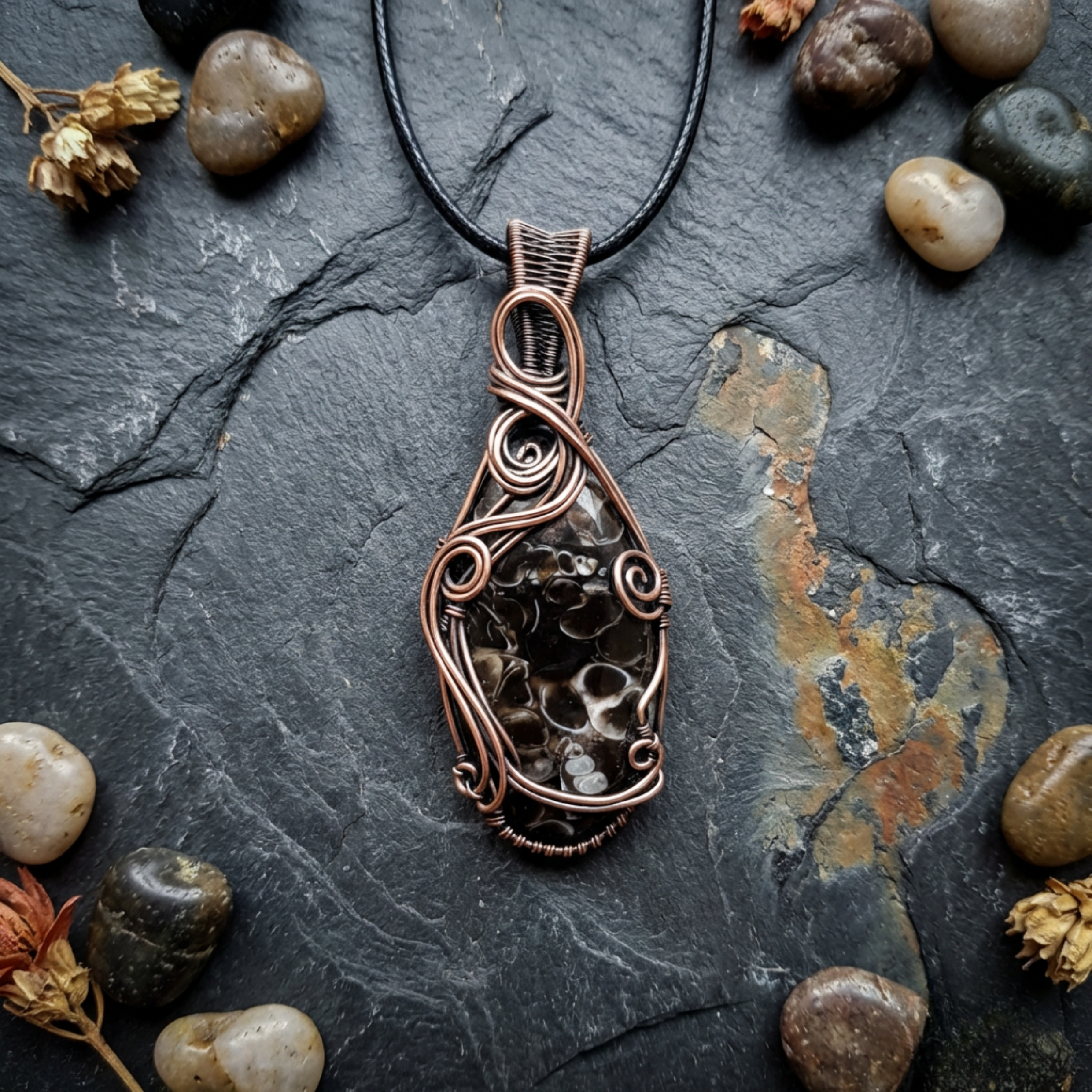 Copper wire-wrapped pendant with embedded stones on a dark stone surface with pebbles and dried plants.