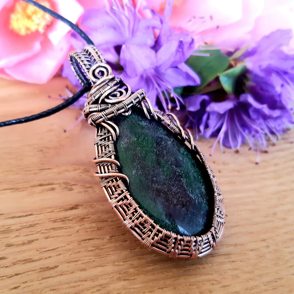 Teardrop-shaped pendant with copper wire wrapping on a wooden surface with purple flowers in the background
