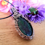Teardrop-shaped pendant with copper wire wrapping on a wooden surface with purple flowers in the background