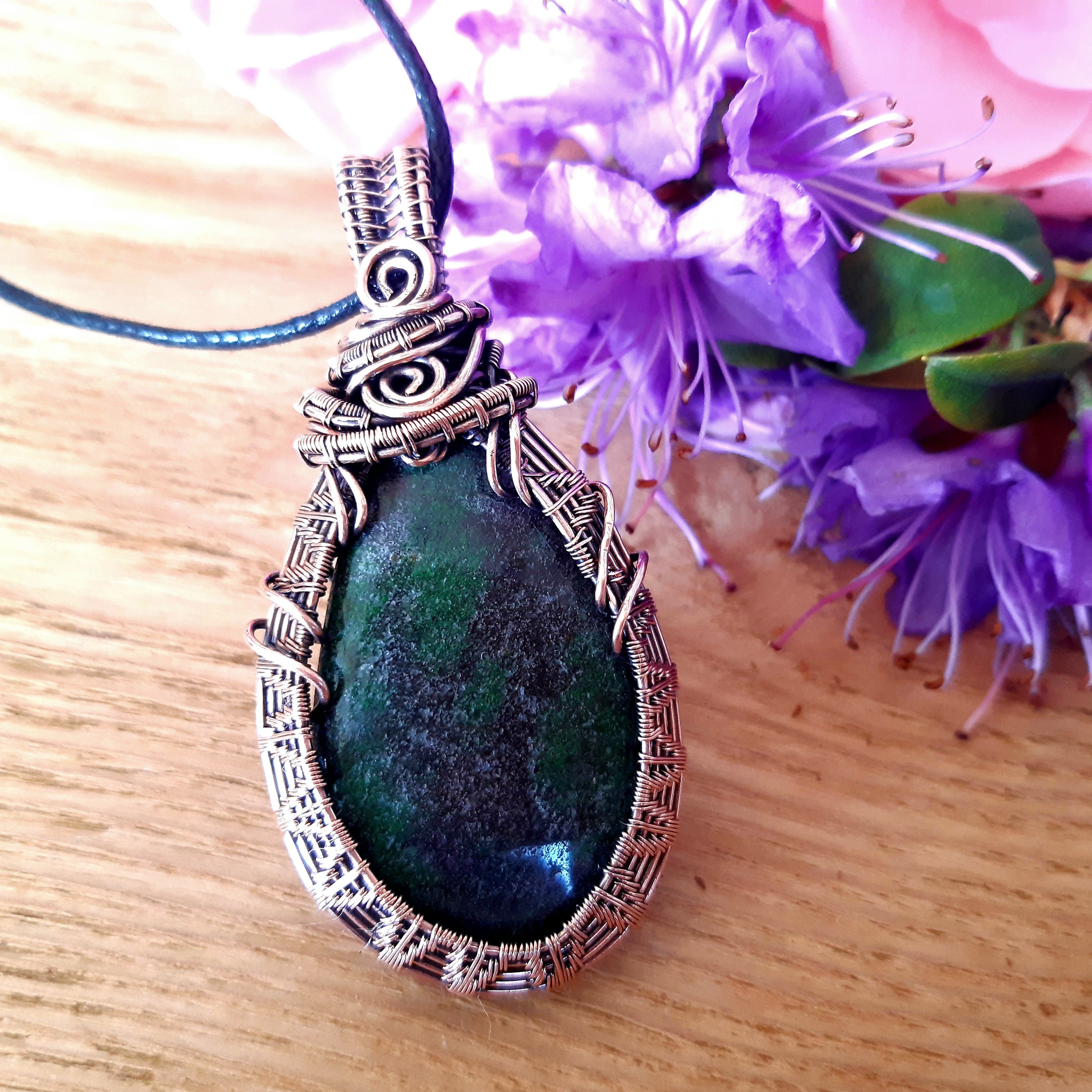 Green stone pendant with copper wire wrapping on a wooden surface with purple flowers in the background
