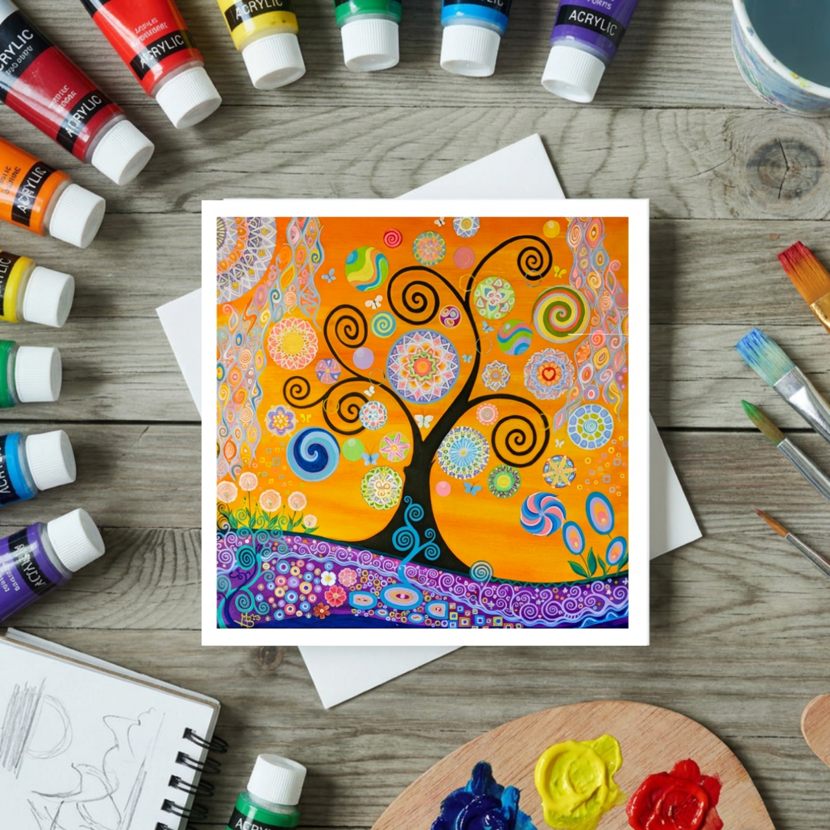 Colourful abstract spiral Tree of Life artwork on a card with acrylic paint bottles and brushes on a wooden surface