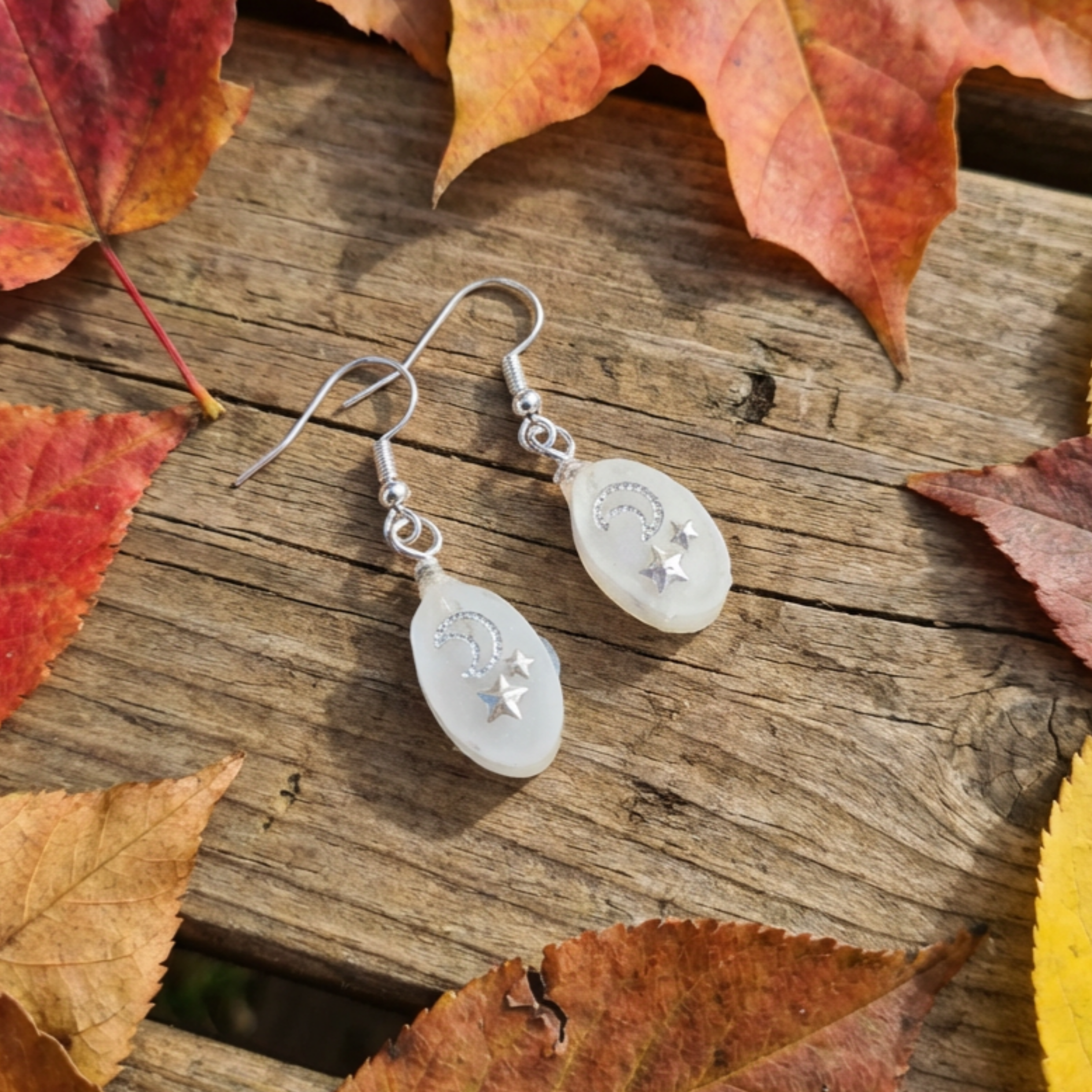 White teardrop earrings with star designs on a wooden surface with autumn leaves.