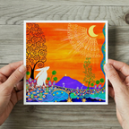 Colourful artwork of a white Moon Gazing hare in a Glastonbury Tor  landscape held by two hands on a wooden surface