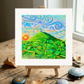 Colourful abstract painting of Glastonbury Tor on a small easel with decorative stones on a wooden surface.