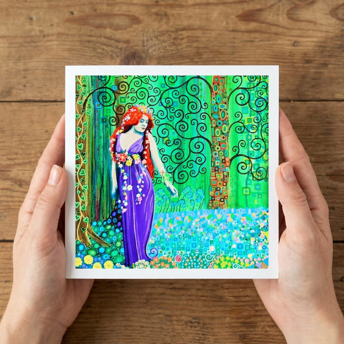 Colourful artwork greeting card of a woman with flowing hair and dress held by hands on a wooden surface