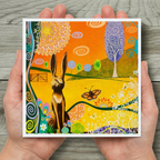 Colourful artwork of a hare and butterfly held in hands on a wooden surface