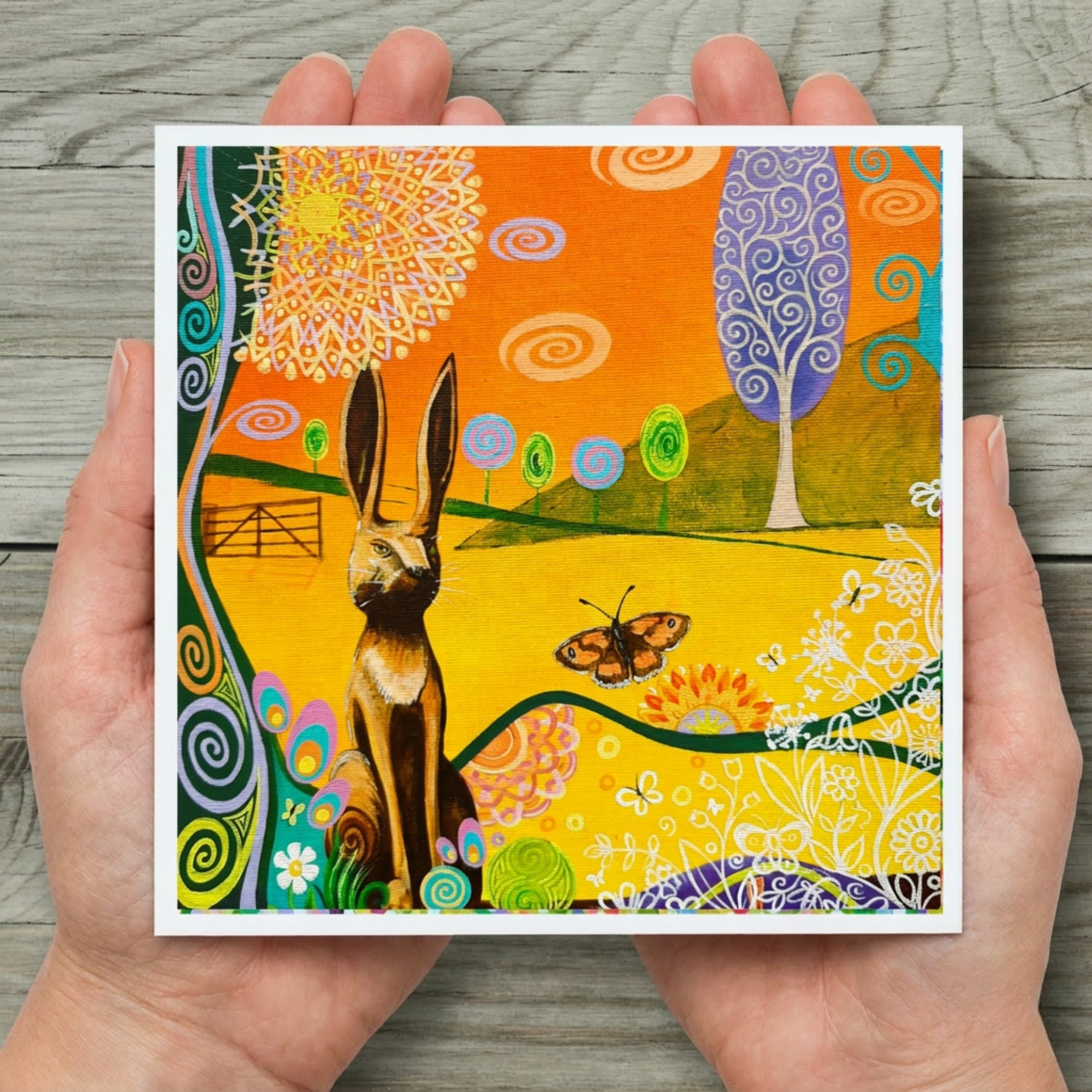 Colourful artwork of a hare and butterfly held in hands on a wooden surface