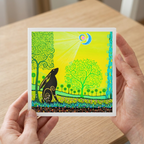 Person holding a colourful card with a stylized illustration of a hare and trees on a wooden surface.