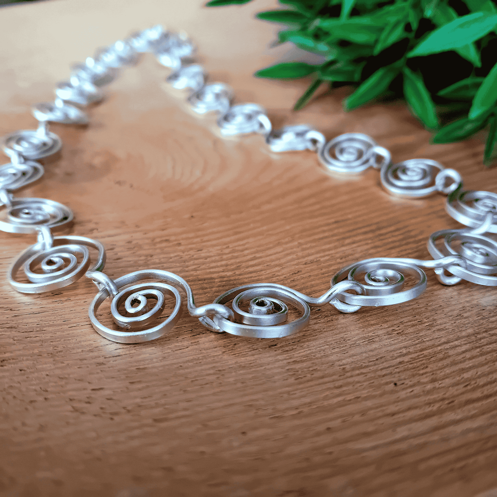 Silver spiral necklace on a wooden surface with green leaves in the background