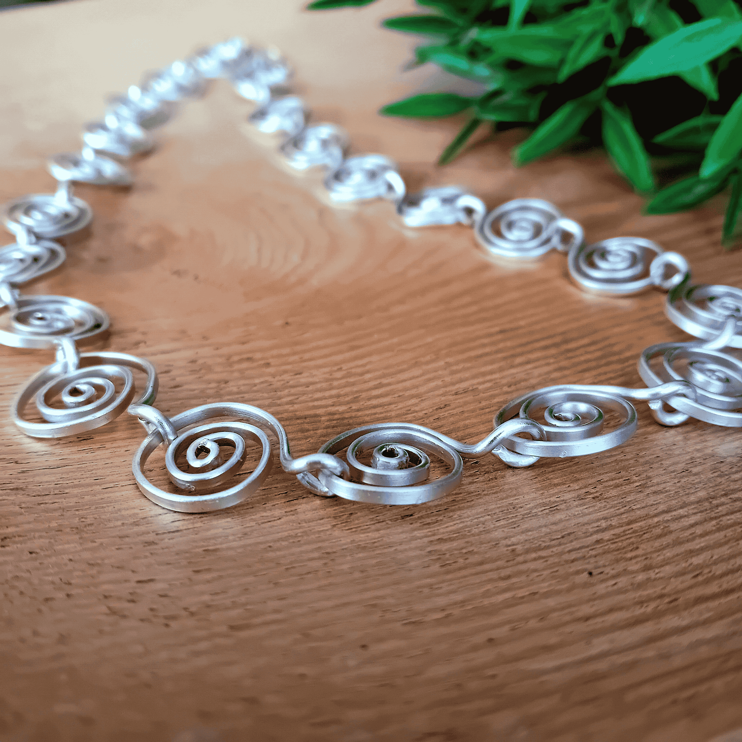 Silver spiral necklace on a wooden surface with green leaves in the background