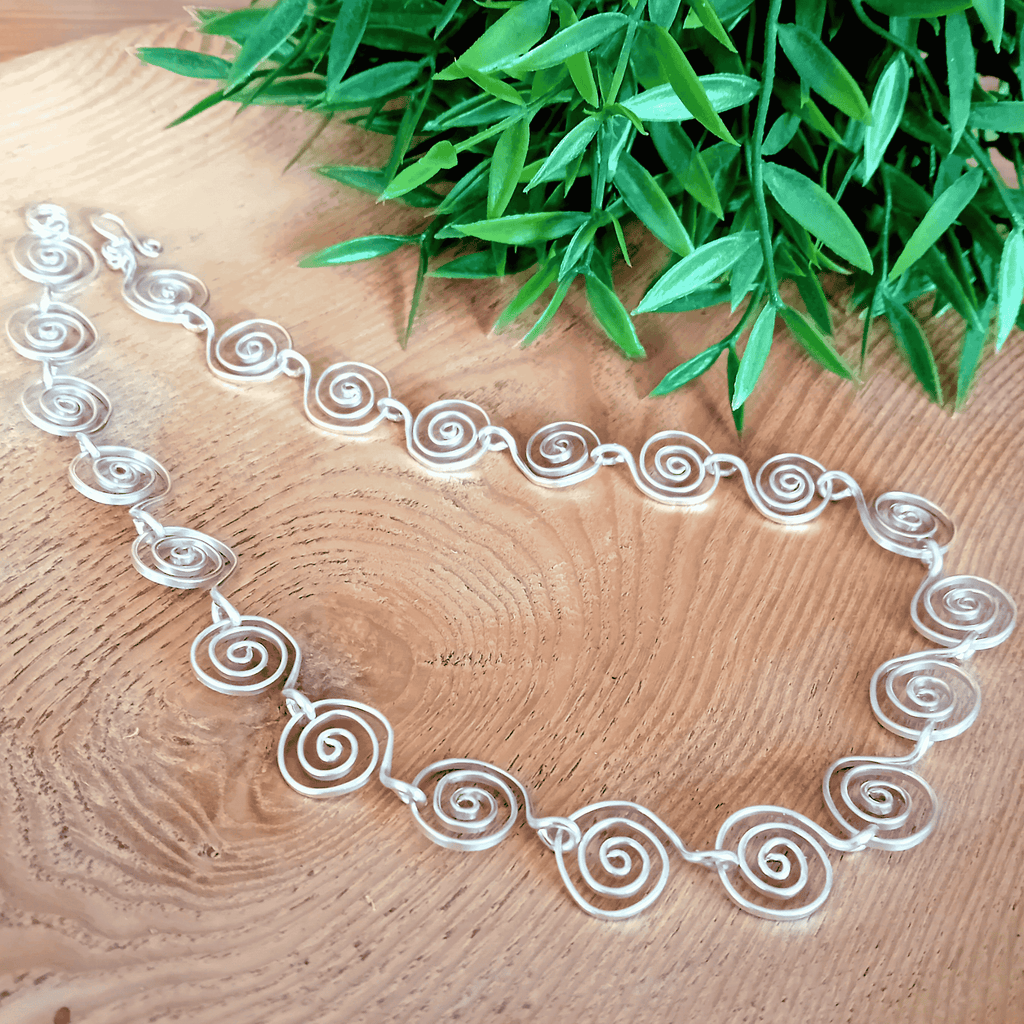 Silver necklace with spiral design on a wooden surface with green leaves in the background