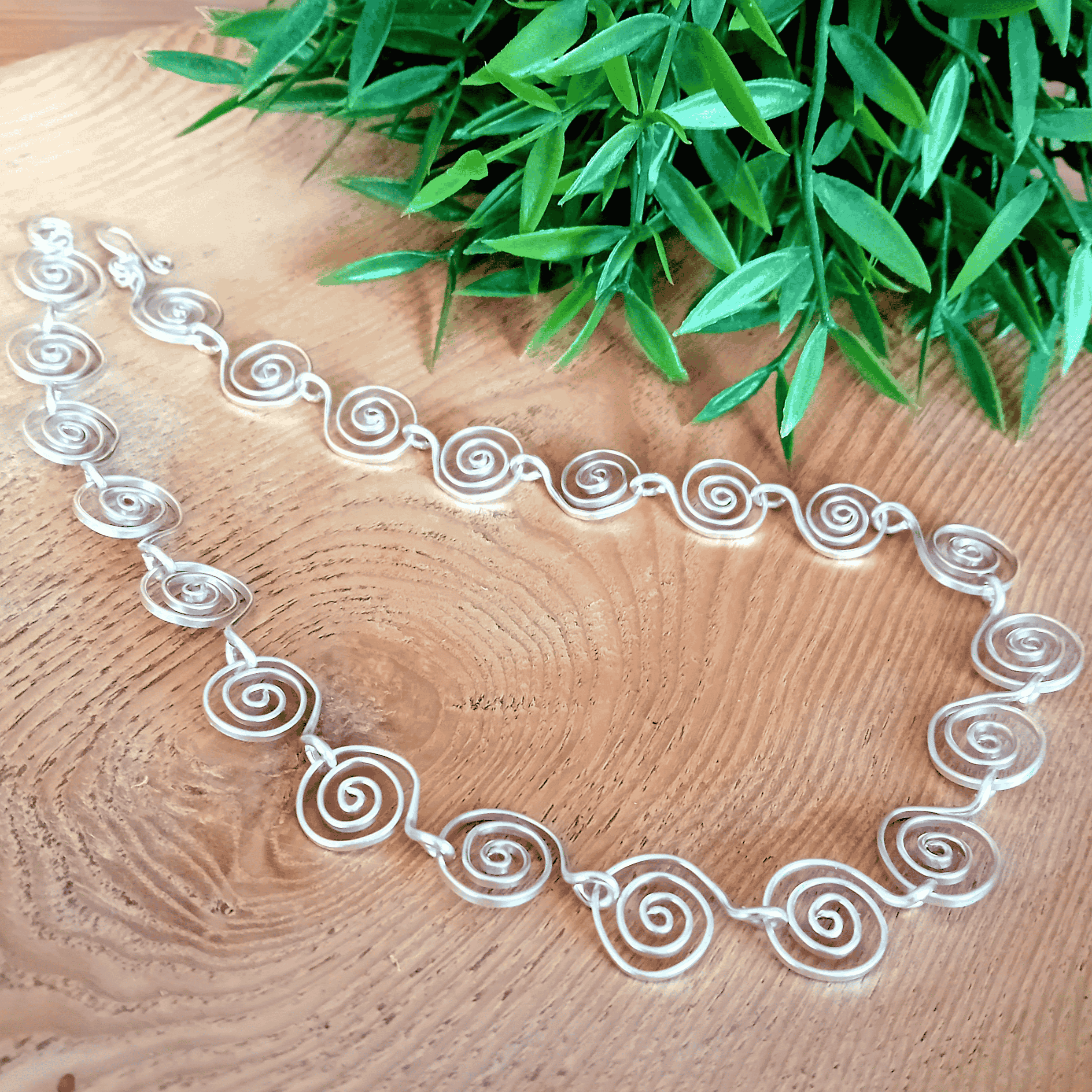 Silver necklace with spiral design on a wooden surface with green leaves in the background