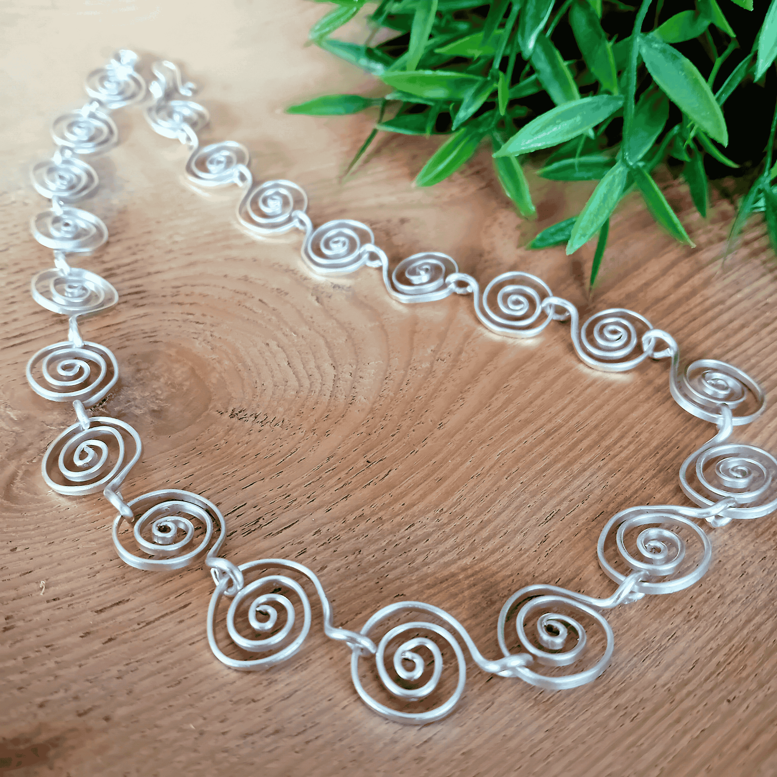 Silver spiral necklace on a wooden surface with green leaves in the background