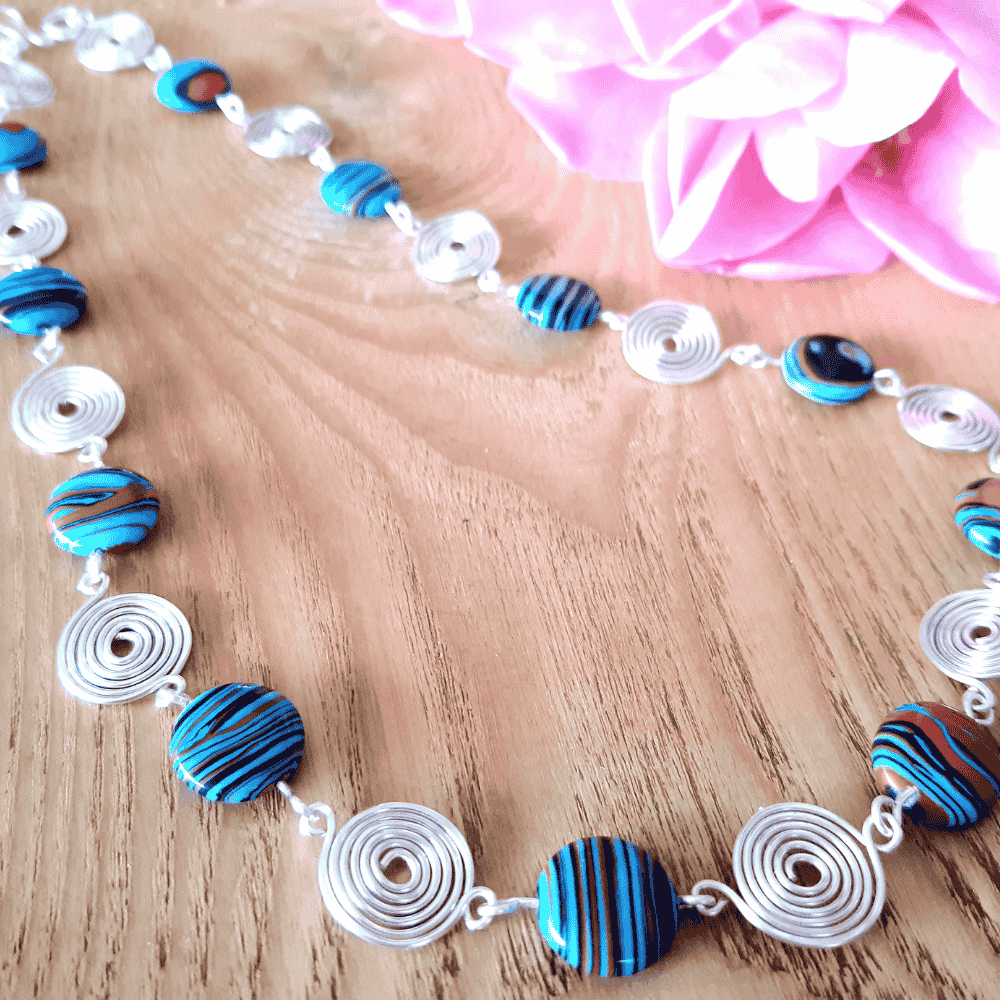 Necklace with blue and black circular beads  and silver spirals on a wooden surface with pink fabric in the background