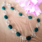 Necklace with green beads and silver spiral accents on a wooden surface with pink flowers.