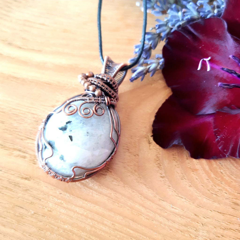 Copper-wrapped pendant with a stone on a wooden surface with a red flower and lavender.