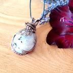 Copper-wrapped pendant with a stone on a wooden surface with a red flower and lavender.